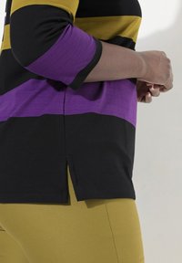 Flerfärgad randig topp i svart, gult och lila material; har trekvartslånga ärmar och sidofickor, ihop med olivgröna byxor.