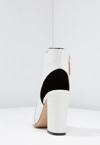 Botín de cuero blanco hasta el tobillo con un panel de ante negro y cremallera dorada. Presenta un tacón grueso blanco y un diseño de punta suave.
