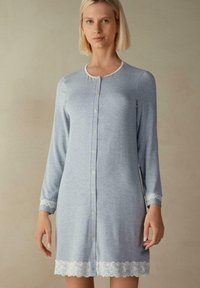 Ljusblå nattklänning med knappar, tillverkad av mjukt tyg, med långa ärmar och vit spetskant vid ärmsluten och nederkanten.