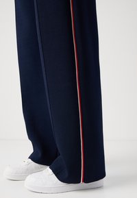 Marineblaue Jogginghose mit roten und weißen Seitenstreifen, aus weichem Stoff. Kombiniert mit weißen, strukturierten Sneakers mit einem Logo.