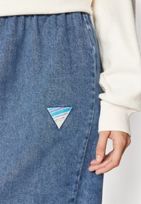 Jupe en denim bleu clair, avec une étiquette triangulaire bleue et blanche à l'avant. Texture douce avec une taille froncée. Chemise blanche à manches longues.