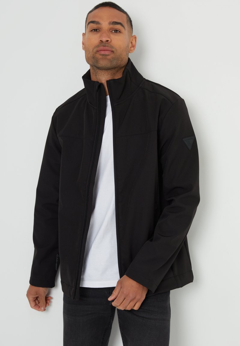 Schwarze Softshelljacke mit hohem Kragen, Reißverschluss vorne und zwei Seitentaschen. Mit einem dezenten Logo auf dem linken Ärmel.