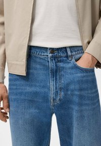 Blauwe denim jeans met zichtbare stiksels, een voorzak en een klassiek pasvormontwerp. Draagt een beige jas en een wit T-shirt.