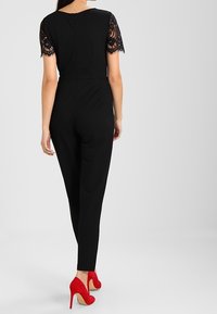 Zwarte jumpsuit met kanten korte mouwen, aansluitend ontwerp en taps toelopende pijpen. Gekleed met felrode hoge hakken.