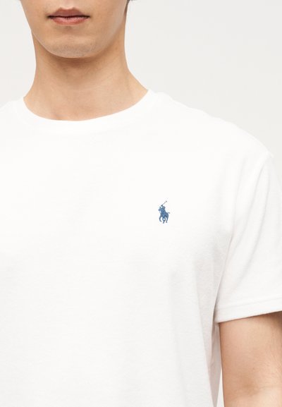 Άνδρας που φοράει λευκό T-shirt με στρογγυλή λαιμόκοψη και μικρό μπλε λογότυπο παίκτη πόλο στο αριστερό μέρος του στήθους, με φόντο απλή επιφάνεια.