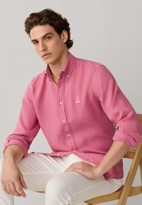 Jeune homme portant une chemise rose boutonnée et un pantalon blanc, assis sur une chaise en bois devant un fond uni.