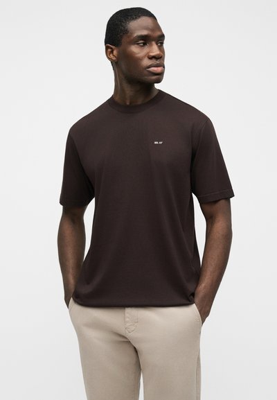 Un homme debout, les mains dans les poches, portant un t-shirt à manches courtes marron foncé avec le texte "NN.07" et un pantalon beige, sur un fond uni.