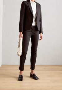 Blazer negro sobre una camisa blanca, combinado con pantalones negros a medida y mocasines negros brillantes. Sosteniendo un bolso blanco estructurado. Suelo de madera clara.