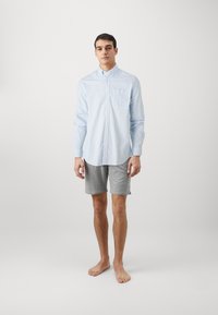 Camisa de botones a rayas azul claro con un bolsillo en el pecho, combinada con pantalones cortos grises. La camisa tiene mangas largas y un dobladillo curvado.