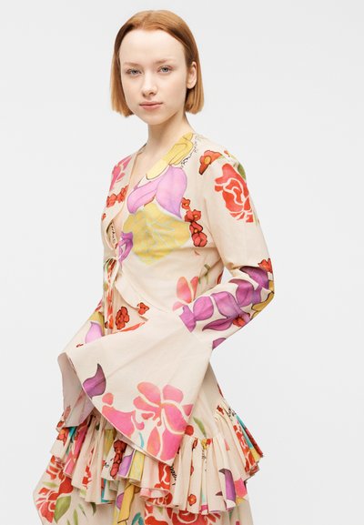 Νέα γυναίκα με κοντά κόκκινα μαλλιά φοράει ένα μακρυμάνικο φόρεμα με λουλούδια, μανίκια καμπάνα και στρωτά βολάν, σε απλό λευκό φόντο.