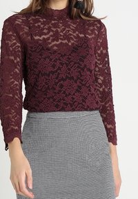 Bordeaux spetsblus med långa ärmar och hög krage, prydd med blommönster. Bärs över en grå rutmönstrad kjol. Genomskinlig textur.