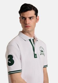 Camisa polo blanca con detalles verdes, que presenta un cuello verde y un logo en el pecho. La manga izquierda muestra un número 3 en verde oscuro.