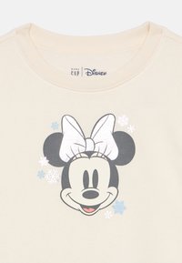 Kremno obarvan pulover z natisnjenim motivom obraza risanega lika z mašno, obdan z grafikami snežink.