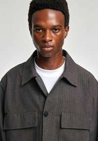 Szara kurtka w paski wykonana z wytrzymałej tkaniny, posiada zapięcie na guziki, dwie przednie kieszenie oraz kołnierzyk. Noszona na białej koszuli.