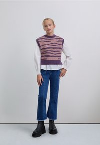 Top sin mangas de punto con rayas moradas y rosas sobre una blusa blanca con volantes, combinado con vaqueros acampanados azules y botas negras gruesas.