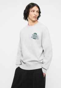 Sudadera gris con cuello redondo, con texto y gráfico en verde en el lado izquierdo del pecho. Tejido suave y puños y dobladillo acanalados.