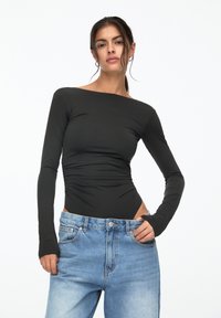 Schwarzer Langarm-Bodysuit mit schulterfreiem Design, versehen mit geraffter Textur an der Seite, kombiniert mit hellblauen, weit geschnittenen Jeans.