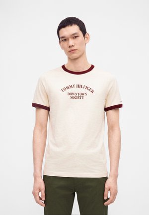 Giovane uomo che indossa una t-shirt color crema con bordino bordeaux e testo "Tommy Hilfiger Downtown Society", abbinata a pantaloni verde scuro.