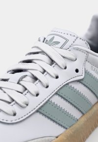 Sneaker bianco in pelle con tre strisce azzurro chiaro. Presenta lacci bianchi piatti, una suola testurizzata e una linguetta con logo vicino alla caviglia.