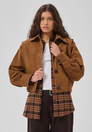 Junge Frau trägt eine braune Wildlederjacke über einem braunen karierten Hemd und weißem Top, steht vor einem schlichten hellen Hintergrund.