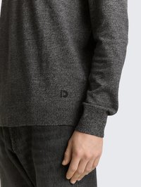 Pilkas megztas megztinis su tekstūruota medžiaga, su ribbiniu apačia ir rankogaliais. Mažas logotipo detailis netoli apatinės apačios; dėvimas su tamsiomis džinsais.