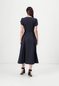 Marineblaues Midi-Kleid mit kurzen Puffärmeln, tailliertem Schnitt und ausgestellt Rock. Glatte Textur und Reißverschluss hinten. Kombiniert mit schwarzen High Heels.