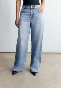 Donna con jeans azzurri a gamba larga, top nero e scarpe appuntite viola scuro, in piedi con una mano in tasca su pavimento in cemento.