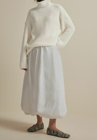 Person, der einen voluminösen weißen Strickpullover, einen lässigen weißen Midirock und flache Schuhe mit schwarz-weißem Zebra-Druck trägt, steht vor einem beigen Hintergrund.
