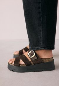 Sandale cu platformă din suede maro, cu o talpă intermediară din plută, talpă exterioară din cauciuc negru și un accent din metal auriu pe cureaua exterioară.