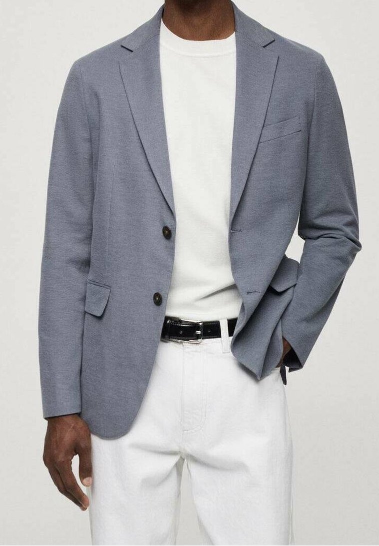 Homme portant un blazer bleu clair, un t-shirt blanc, un pantalon blanc et une ceinture noire, une main dans la poche devant un fond uni.