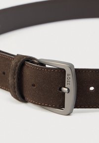 Καφέ σουέντ ζώνη με αγκράφα από ασημί μέταλλο με ανάγλυφο λογότυπο, ραφές λεπτομέρειας και πολλαπλές τρύπες για ρύθμιση.