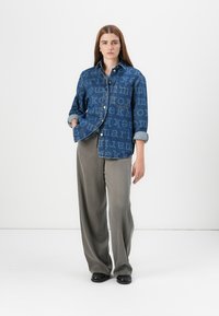 Denimjakke med blå vask og gjentatt tekstmønster, kombinert med løse, grå vide bukser og svarte sko.