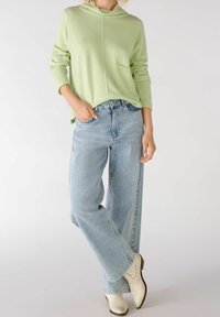 Persona che indossa una felpa verde chiaro, jeans a gamba larga blu chiaro e stivaletti bianchi, in piedi con una gamba incrociata sopra l'altra.