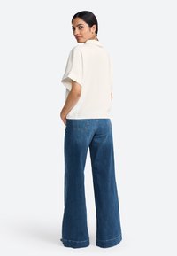 Haut collier à manches courtes crème avec une coupe décontractée, associé à un jean en denim bleu clair à jambes larges, présentant des poches arrière visibles et des détails au niveau de l'ourlet.