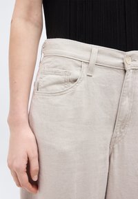 Δεξί χέρι που ακουμπάει δίπλα σε ανοιχτό μπεζ παντελόνι από τζιν με μπροστινή τσέπη και θηλιές για ζώνη, σε συνδυασμό με μαύρο ριμπ τοπ.