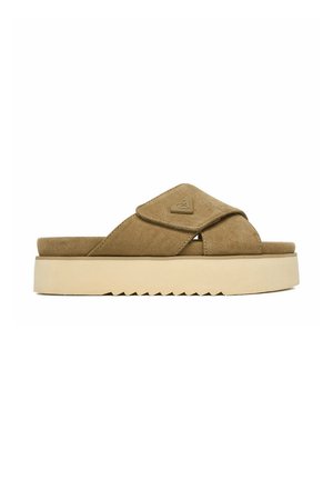 Bruine suède instapsandaal met dikke beige plateauzool en gekruiste bandjes, met een klein driehoekig logo op het bovenste bandje.