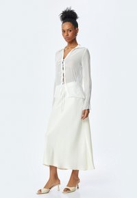 Blouse blanche plissée avec détail de lien à l'avant, associée à une longue jupe blanche lisse et des mules à talons beiges, sur fond clair.