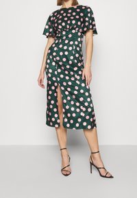 Grön satin klänning med rosa prickar, korta fladdrande ärmar och en sidoklyfta. Har en figursydd midja och en midilängdsslut. Svarta klacksandaler.