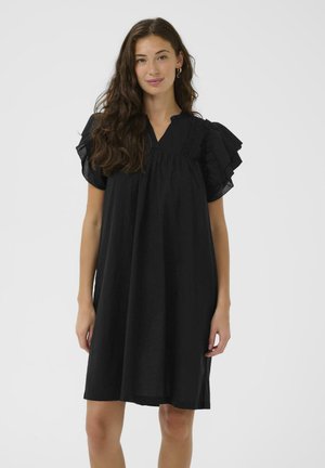 Žena s dlhými tmavými vlasmi v voľných čiernych šatách do kolien s volánovými krátkymi rukávmi na jednoduchom bielom pozadí.