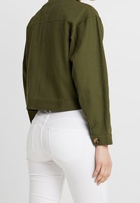 Veste cropped vert olive au design classique, dotée de longues manches et d'un détail de bouton sur les poignets. Portée sur un jean blanc.