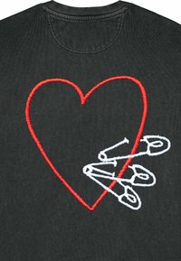 Camiseta negra con un gran corazón bordado rojo y tres alfileres de seguridad blancos, situados debajo del diseño del corazón.
