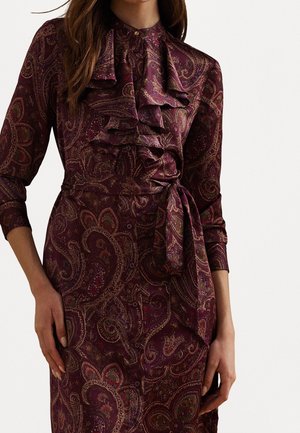 Vrouw draagt een paars paisleypatroon jurk met lange mouwen, knoopsluiting aan de voorkant, ruches op de borst en een strikriem in de taille, tegen een effen achtergrond.
