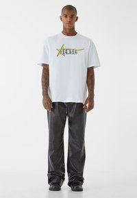 Balta didelė T-shirt su geltonu žvaigždės grafiku ir užrašu "SENSE.", derinama su blizgiais juodais dirbtinės odos kelnėmis ir juodais batais.