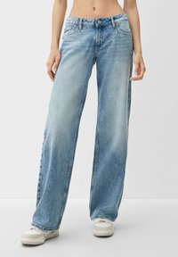 Mujer vestida con jeans descoloridos de un azul claro y zapatillas blancas, de pie contra un fondo liso de color gris claro.