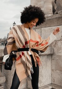 Châle rayé beige et noir avec des franges, porté ceinturé à la taille. Associé à un pull blanc et un legging noir. Petit sac à main noir.