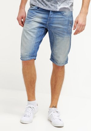 Pantalones cortos de mezclilla en azul claro con una textura desgastada. Presentan dobladillos enrollados, bolsillos frontales y un bolsillo lateral adicional. Combinados con zapatillas blancas.