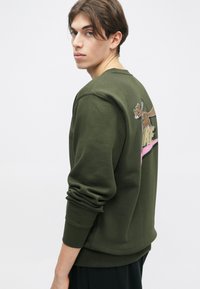 Sweatshirt verde-oliva feito de mistura de algodão, com um gráfico impresso nas costas que apresenta um personagem de cartoons e texto em destaque. Design de corte slim.