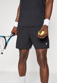 Black sports shorts made of lightweight fabric, featuring a small logo on the side, held by a person with a tennis racket and ball.