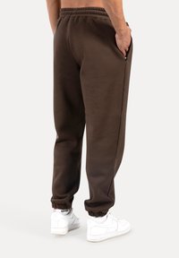 Brązowe spodnie dresowe wykonane z miękkiego materiału, o luźnym kroju, z elastycznym pasem, bocznymi kieszeniami i elastycznymi zakończeniami nogawek, zestawione z białymi butami sportowymi.