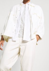 Witte oversized linnen jas met zilveren drukkers over een wit overhemd en lichtgrijze T-shirt, gecombineerd met off-white broek. Soepele textuur.